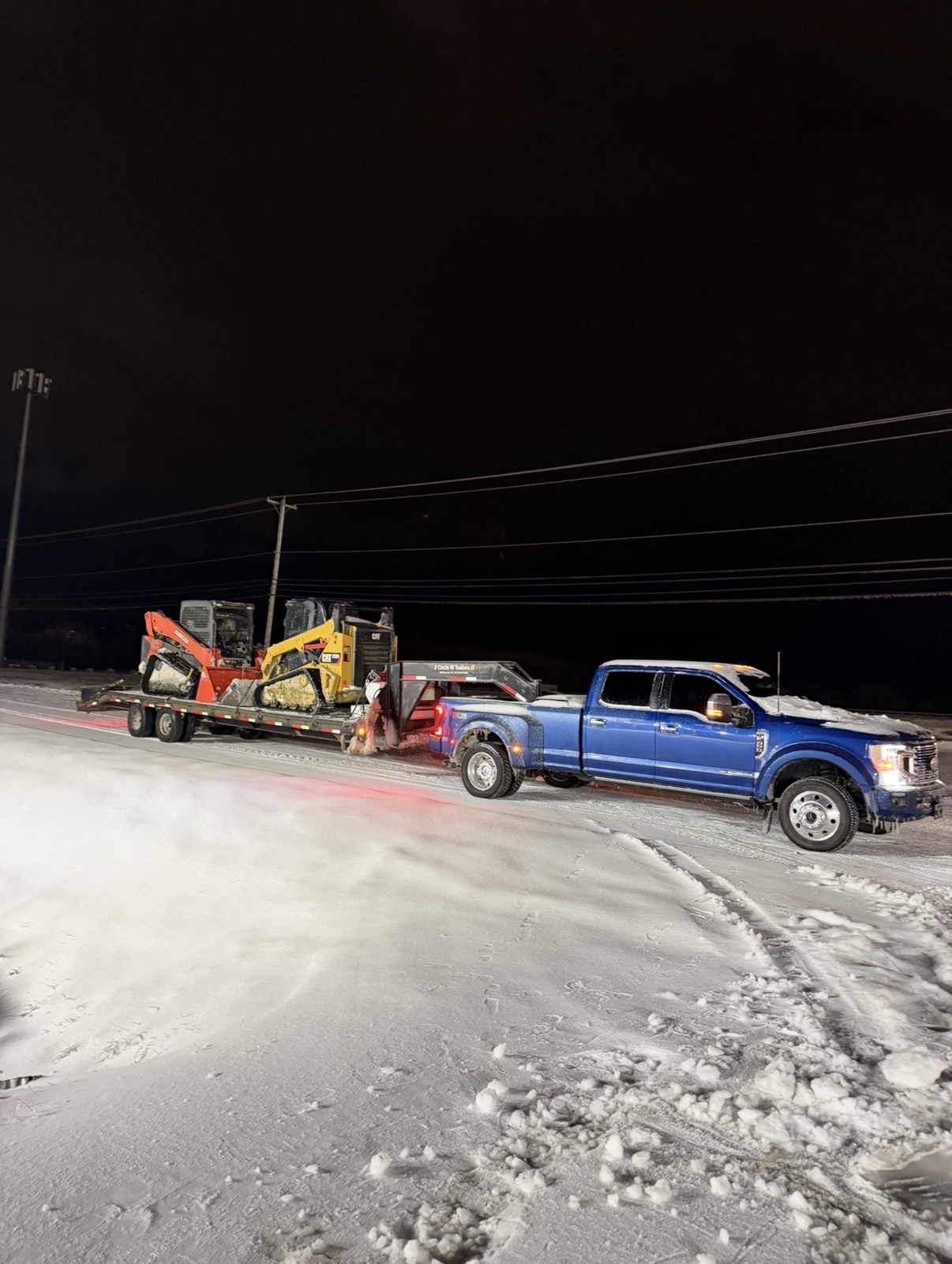 Blue truck hauling two skid steers on a snowy night